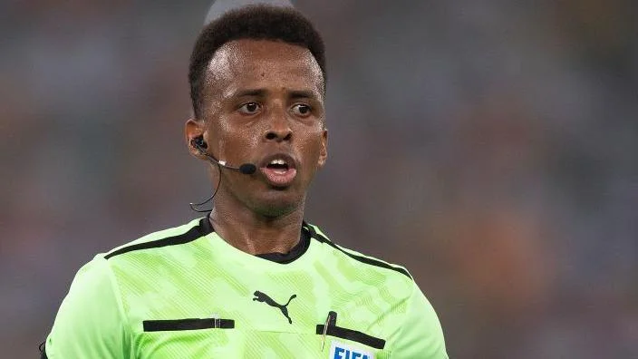 Omar Artan refereeing a match, with earpiece and microphone, and a Fifa badge on his light green shirt