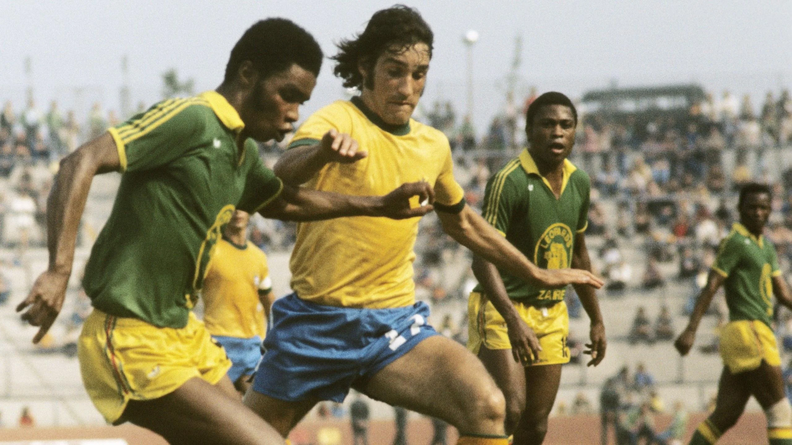An image from the 1974 World Cup in West Germany, with Zaire taking on Brazil. The central part of the image shows a Zaire player, wearing yellow shorts and a green shirt with yellow trim, tussling for a ball which is out of shot below the base of the image, against a Brazilian opponent who is wearing blue shorts and a yellow shirt. The Zaire player is pushing out his left arm at chest level in a bid to ward off the tackle. Two more Zaire players are watching on in the background, while behind them is a sparsely populated concrete stand