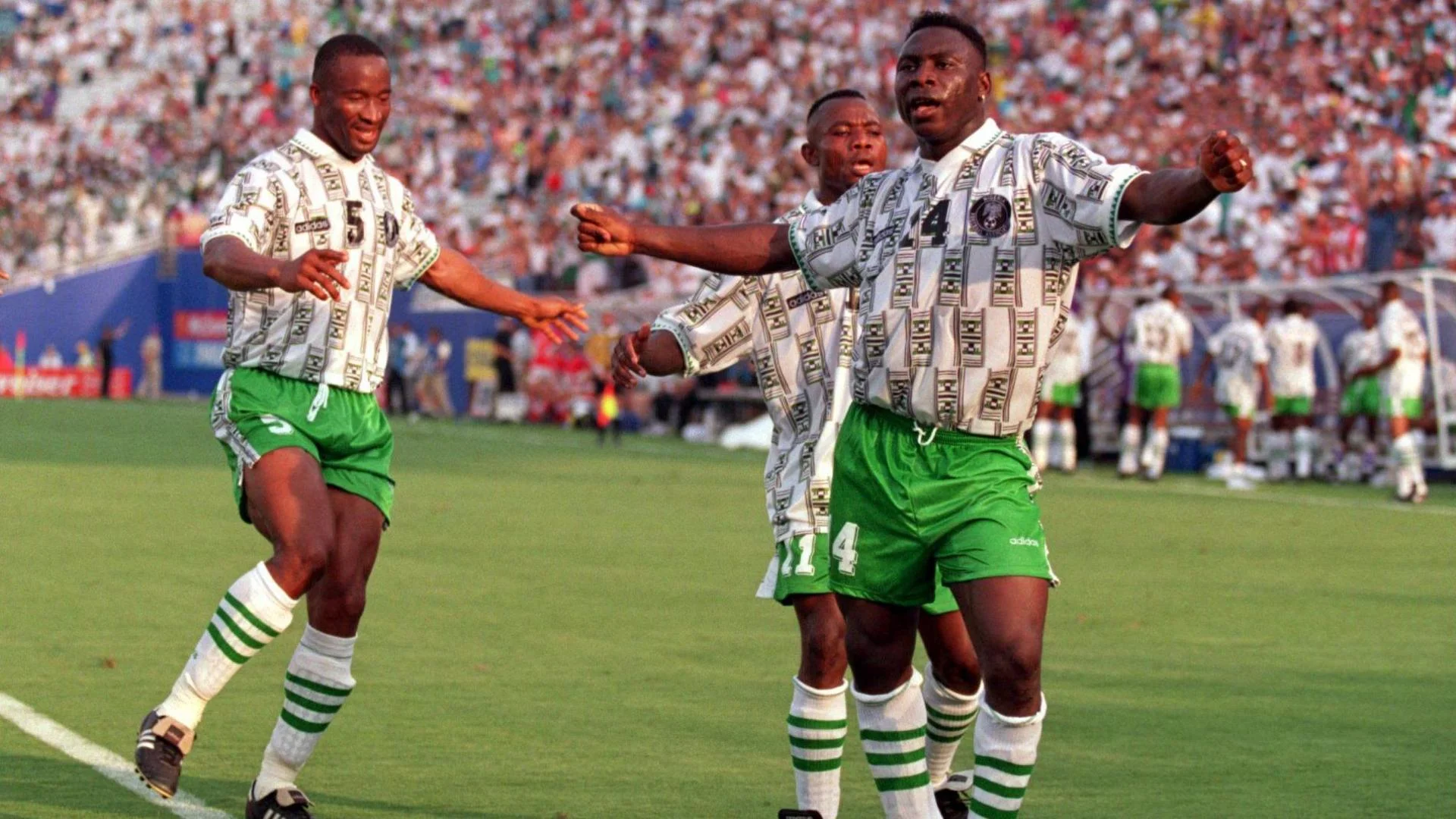 An image from the 1994 World Cup in the USA shows three Nigeria players celebrating a goal. Scorer Daniel Amokachi dances with movements that suggest a wiggling of the hips, with his arms spread wide, clicking his fingers, and with one straight leg and another tilted at the knee. One of the other players is directly behind Amokachi and preparing to wrap his arms around him, while the other is approaching from the left-hand side of the image, also with arms spread and a big smile. All three players wear white socks with three green horizontal stripes on the upper portion, green shorts, and a white shirt dominated by an intricate grey and black pattern which is broken into alternating blocks, giving off a traditional African vibe. A stand containing a packed crowd is blurred in the background, as is the Nigerian dugout, complete with a number of players on their feet